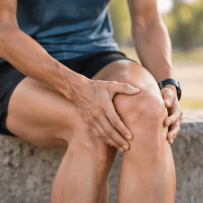 A seated runner holds the front and sides of the knee while experiencing discomfort associated with conditions addressed by patellofemoral pain syndrome exercises