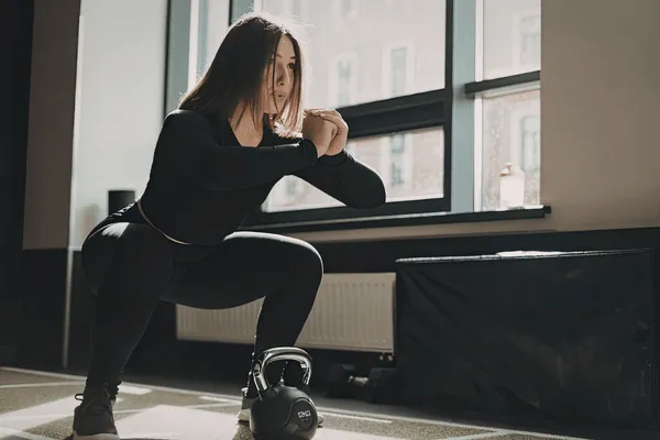 Woman holds deep squat in gym near kettlebell, practicing functional movement training for strength