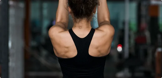 Rear view of an athlete in a black racerback top performing an overhead arm raise inside a sports rehab facility.