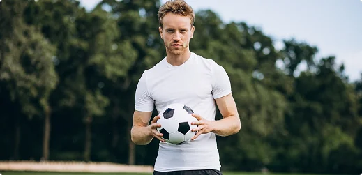 Soccer player holding a ball outdoors in front of a tree line.